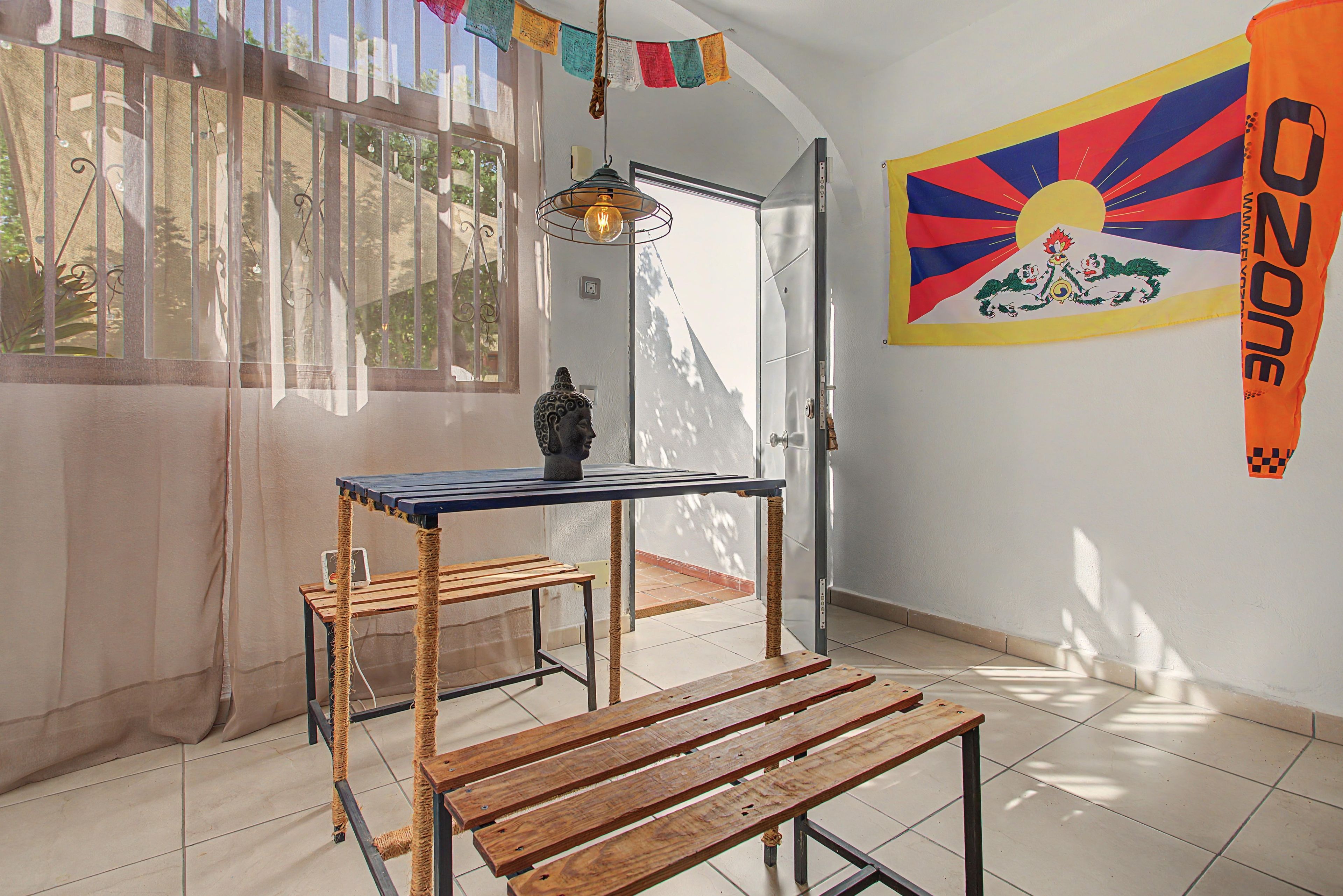 Ein Zimmer mit einer Holzbank, einem Tisch mit einer Lampe, einer Wand mit einer bunten Flagge und einer Tür, die nach draußen führt.