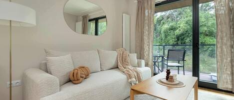 A modern living room with a sofa, a coffee table, a lamp, and a view of greenery through a large window.