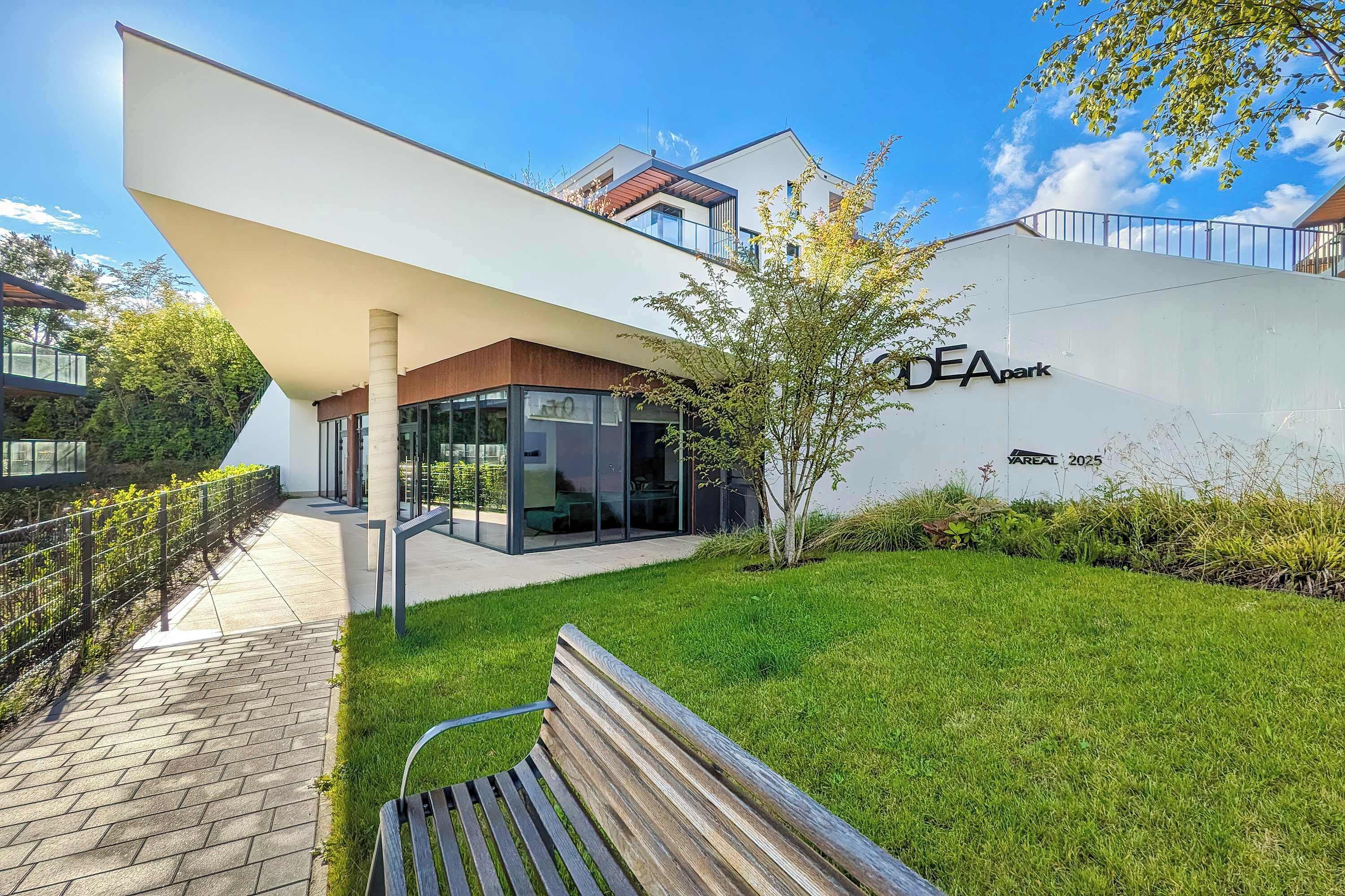 Een modern gebouw met een glazen gevel, een gazon met een bankje en een bordje met de tekst 'IDEApark'.