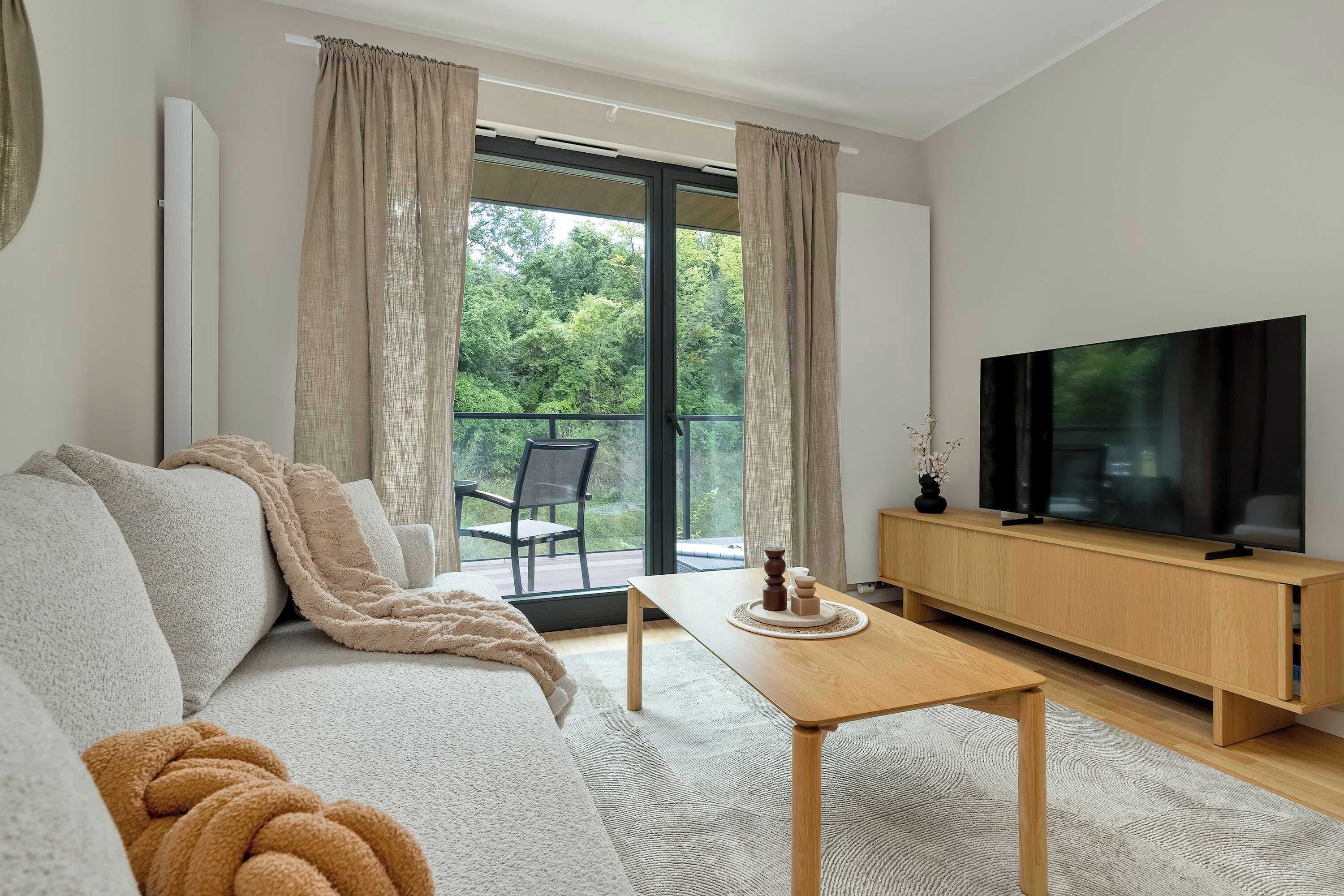 Ett modernt vardagsrum med en soffa, ett soffbord, en tv på ett träställ och utsikt över grönskan genom fönstret.
