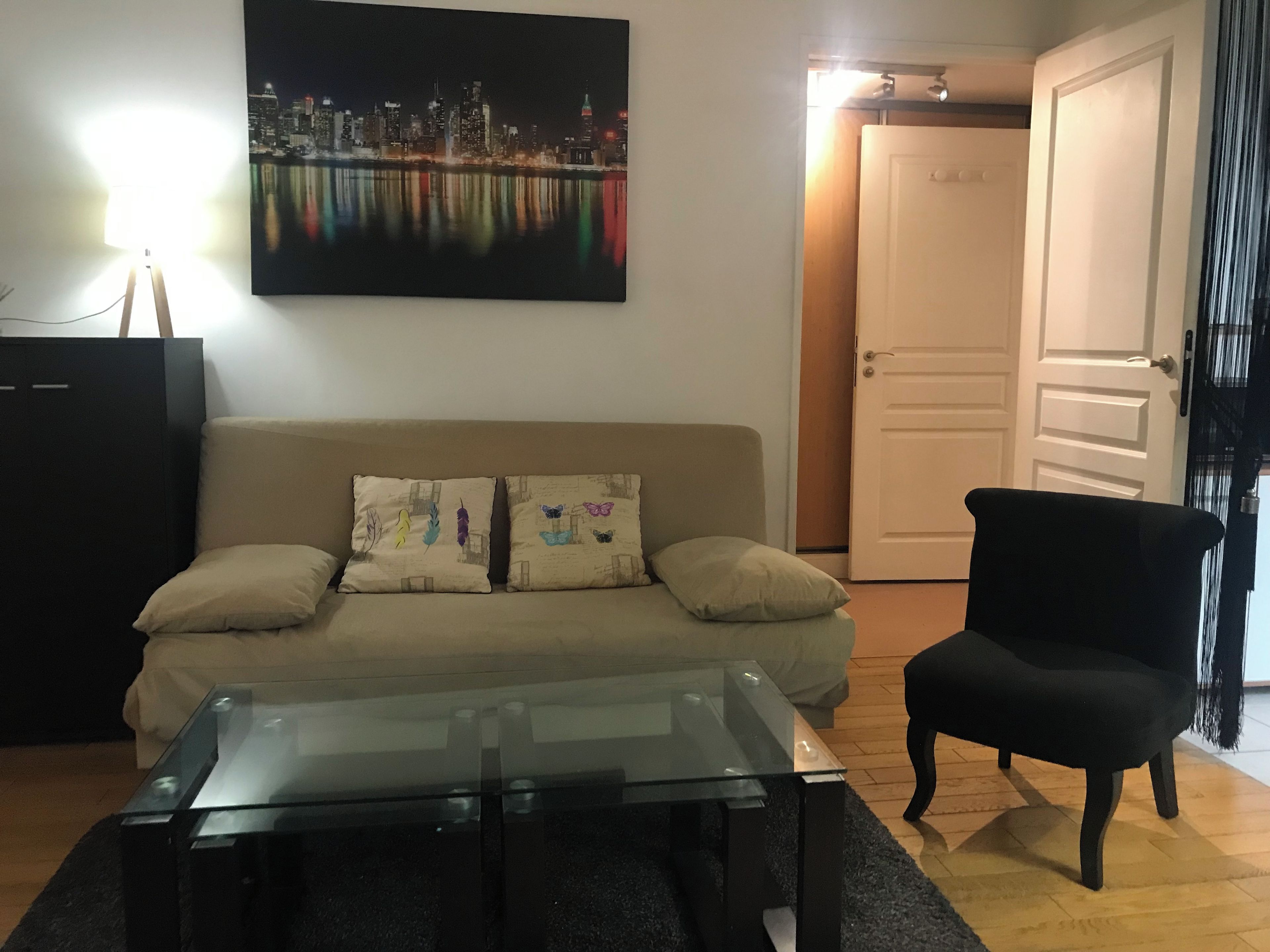 A living room with a sofa, a glass coffee table, a black armchair, and a cityscape painting on the wall.