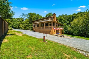 A beautifully crafted log cabin sits nestled among lush summer greenery, offering a peaceful retreat surrounded by mature trees and natural landscape. - Douglas Lakefront Cabin, Boat Ramp, Private Dock, Hot Tub! (Sevierville)