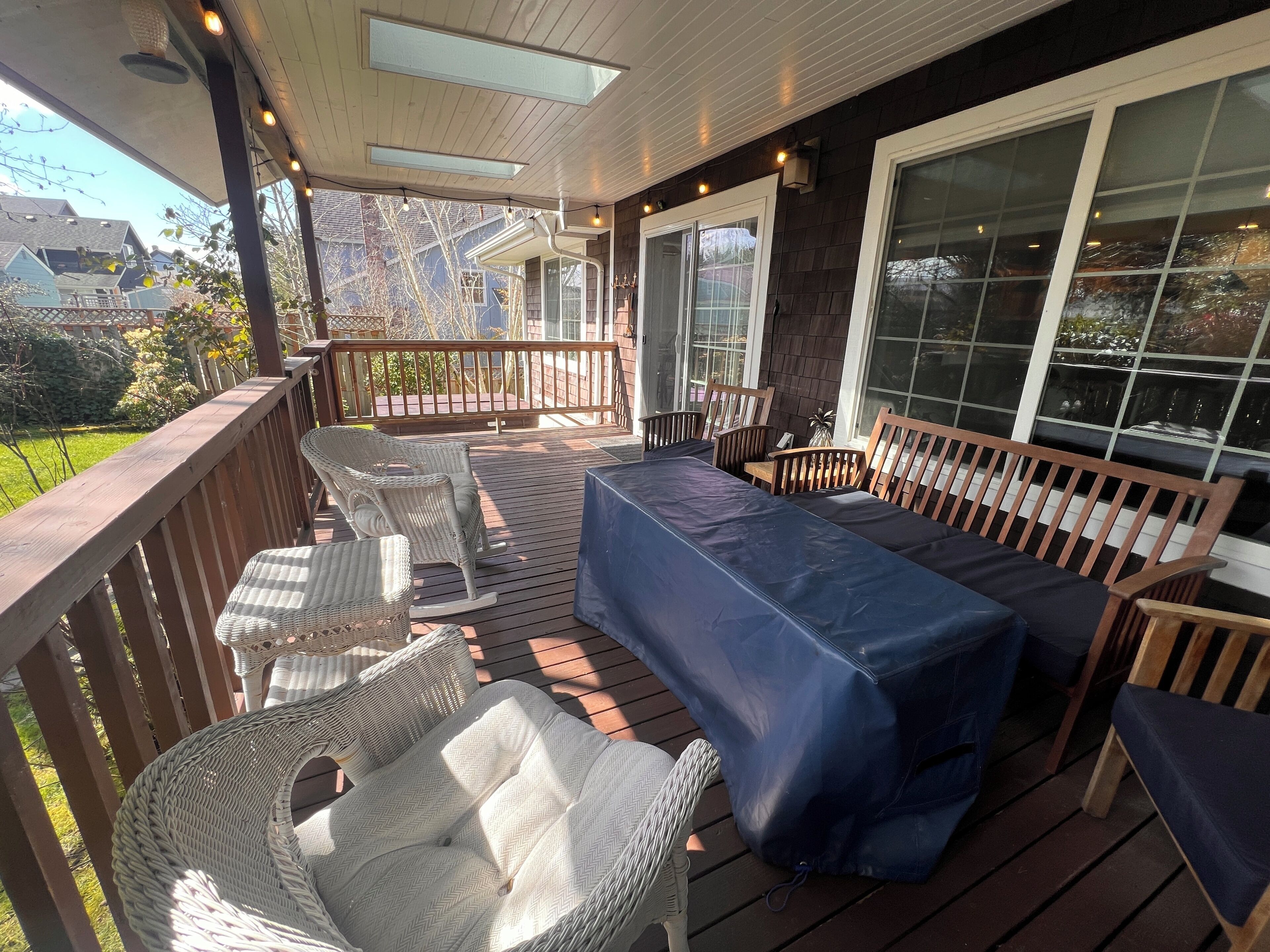 Une terrasse extérieure couverte, aménagée avec des meubles en osier, un banc bleu avec coussins et de grandes fenêtres.