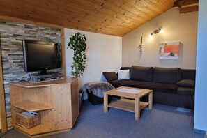 Ein gemütliches Wohnzimmer mit Holzbalkendecke, einem Flachbildfernseher, einem dunklen Sofa, einem kleinen Holztisch und einer Topfpflanze.