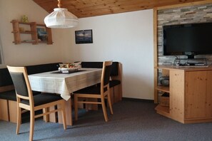 Ein Essbereich mit einem Tisch für vier Personen, Holzstühlen, einem Fernseher auf einem Holzschrank und einer Decke mit freiliegenden Balken.