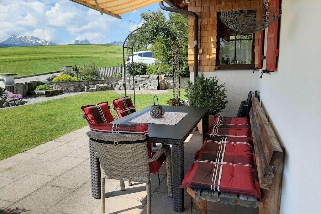 Een terras buiten met een tafel, stoelen, een hanglamp en uitzicht op een groen landschap.