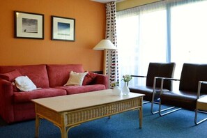 A room with a red sofa, a small table, two framed pictures on the wall, and a window with curtains. - Apartment with 1 bedroom (Hollum Ameland)