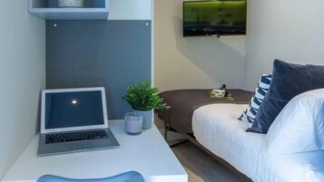 Un dormitorio moderno con cama, un escritorio con una computadora portátil, un televisor montado en la pared y una pequeña planta en el escritorio.
