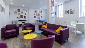 A modern lounge area with purple sofas, yellow and blue round tables, and a ping pong table.