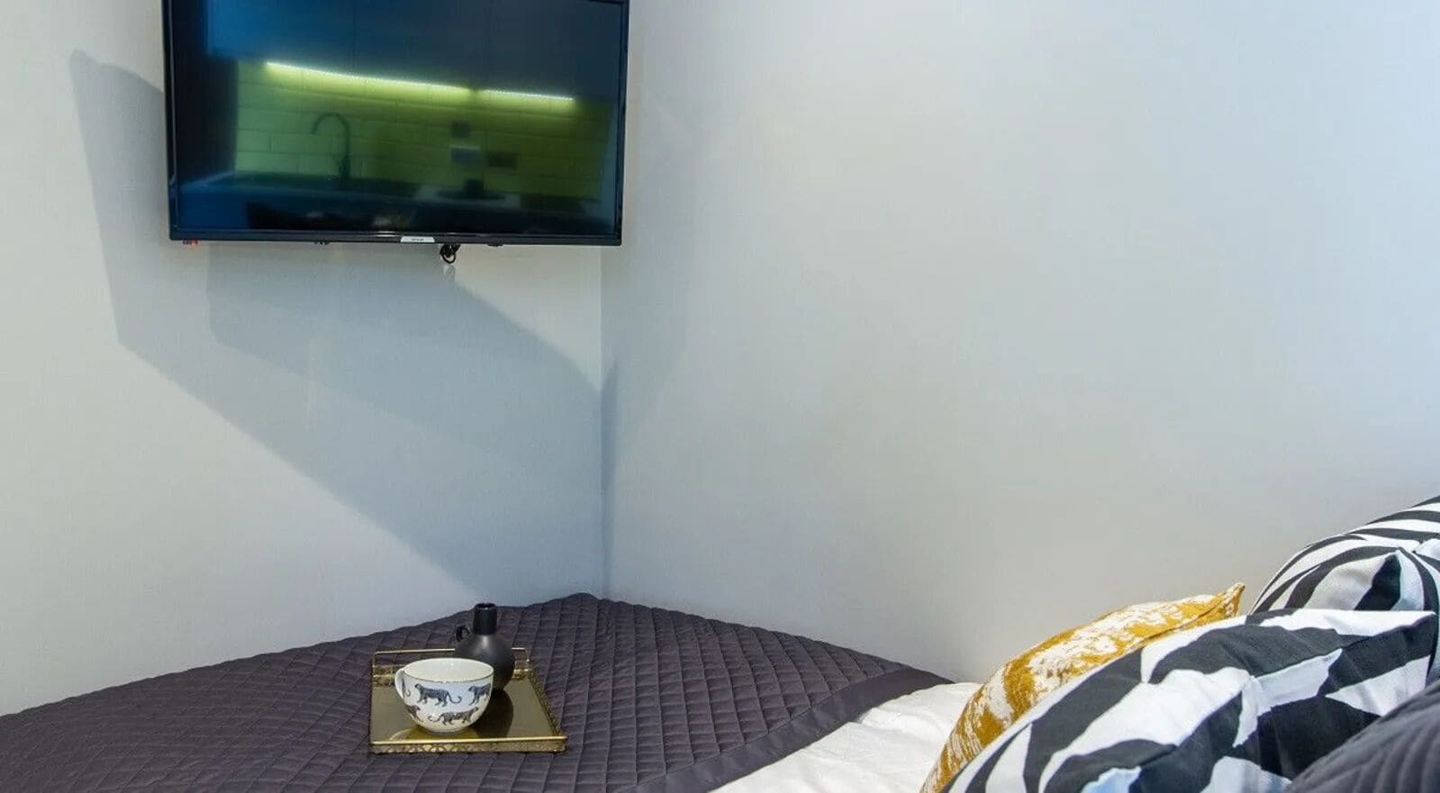 A hotel room with a bed, a flat-screen TV mounted on the wall, and a bedside table with a cup and a small vase.