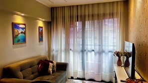 A living room with a sofa, two framed paintings, a large window with sheer curtains, and a flat-screen TV on a stand. - 1904 Nazaré Apto 2 quartos com Garagem (Belém)