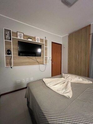 Un dormitorio con cama, armario, televisor montado en la pared y un pequeño estante con adornos.