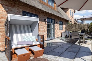 Eine Terrasse mit einem Gebäude mit Reetdach, einem gestreiften Liegestuhl und einem Tisch mit Stühlen.