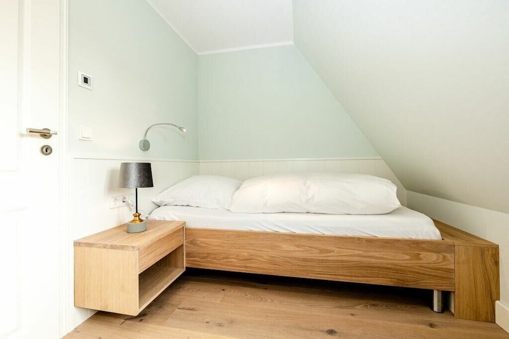 A bedroom with a sloped ceiling, a wooden bed frame, a bedside table with a lamp, and a wall-mounted light fixture.