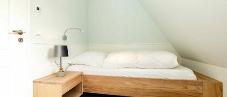 Ein Schlafzimmer mit schrägen Wänden, einem Bettgestell aus Holz, einem Nachttisch mit Lampe und einer Wandleuchte.