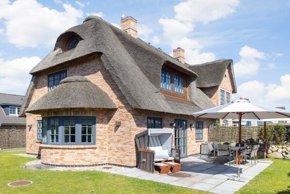 Ein Haus mit einem Reetdach und einer Ziegelaußenfassade, eine Terrasse mit Gartenmöbeln und eine Steinmauer im Hintergrund.