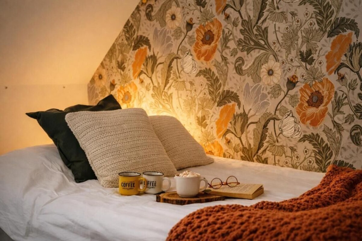 Un llit amb paper pintat floral, dos coixins, dues tasses de cafè, un llibre i una parella de gaussets.