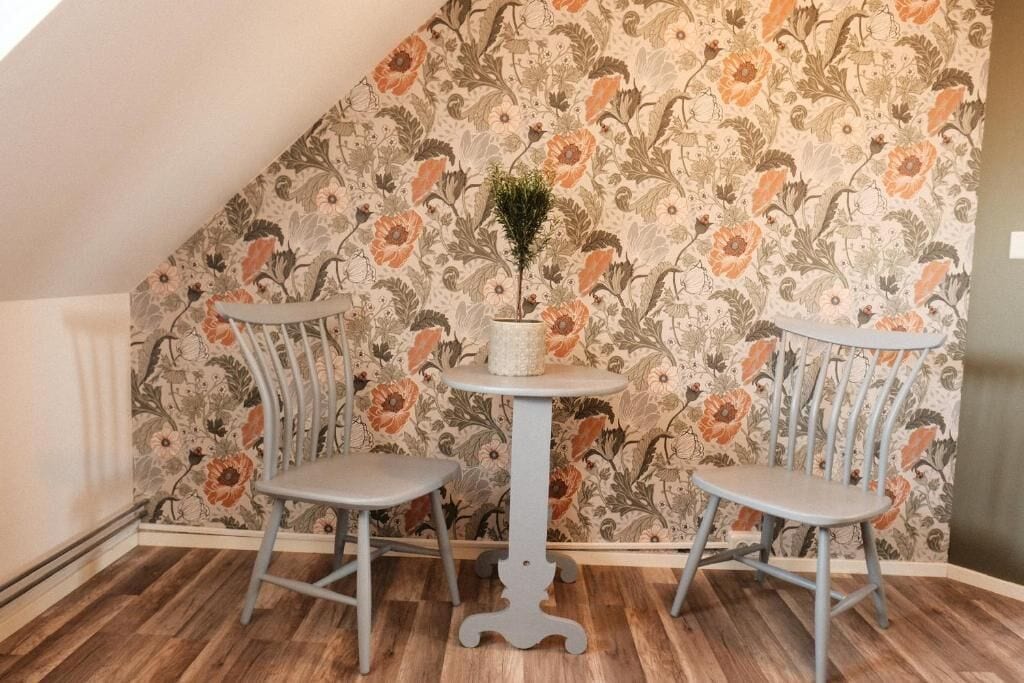 Een kamer met een schuine balklaag, twee grijze stoelen, een klein, rond tafeltje met een bloempot en een behang met een bloemenpatroon.