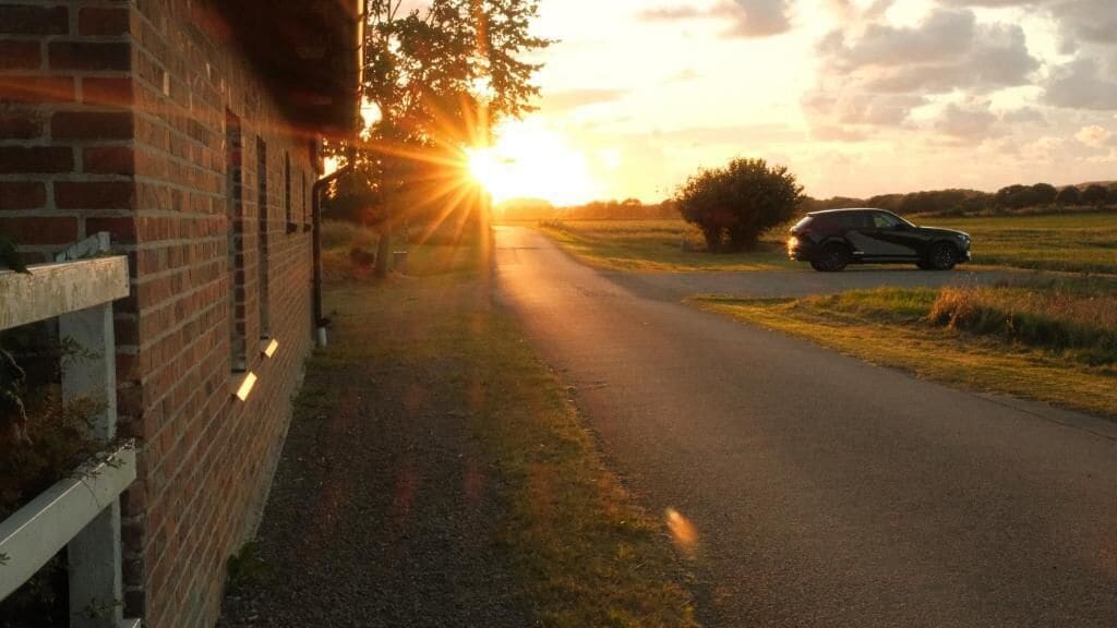 A rural road with a brick building on the left, a parked car on the right, and a sunset in the background.