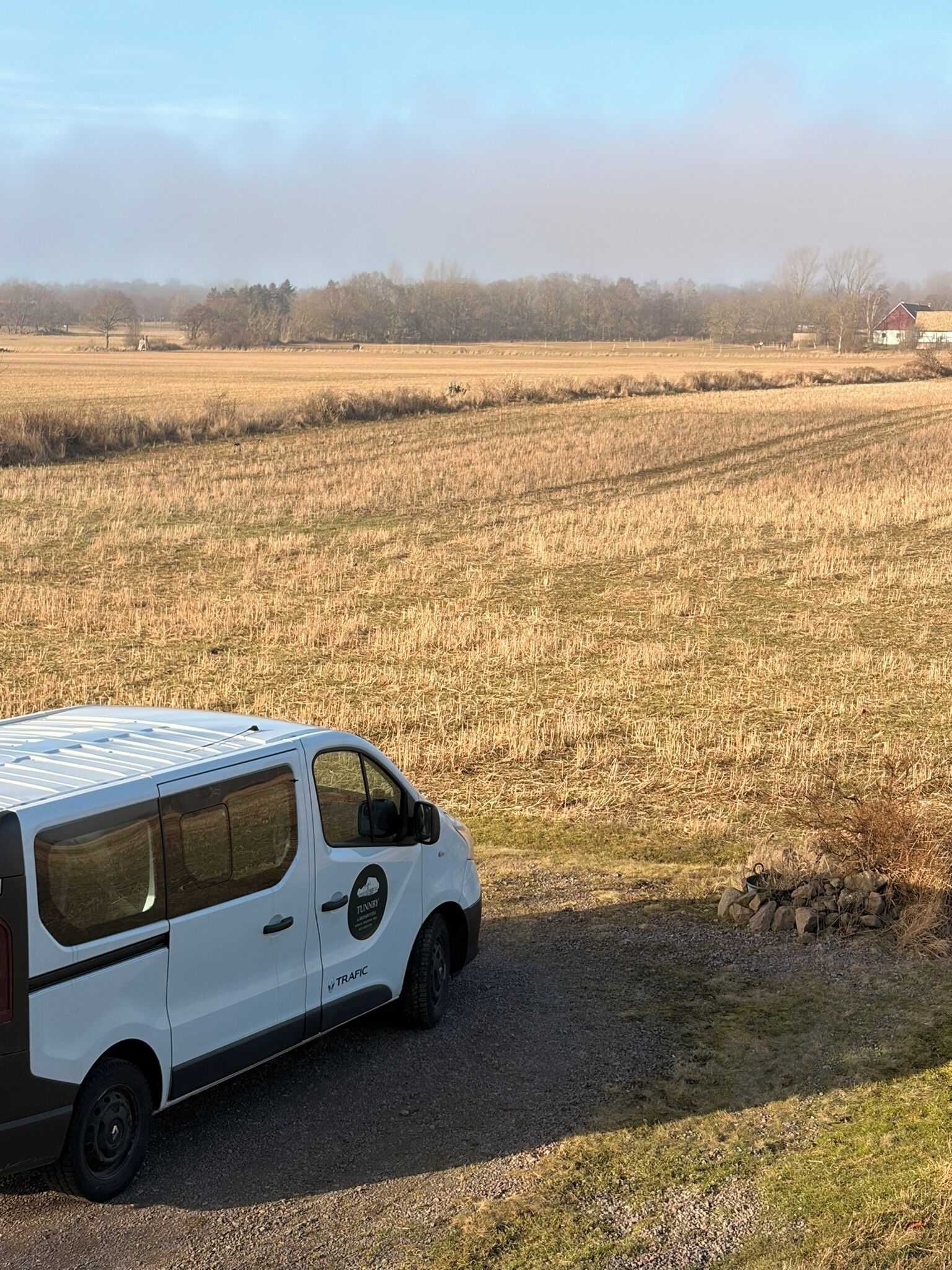 Uma van branca estacionada em um caminho de cascalho em um campo rural, com um celeiro ao fundo.