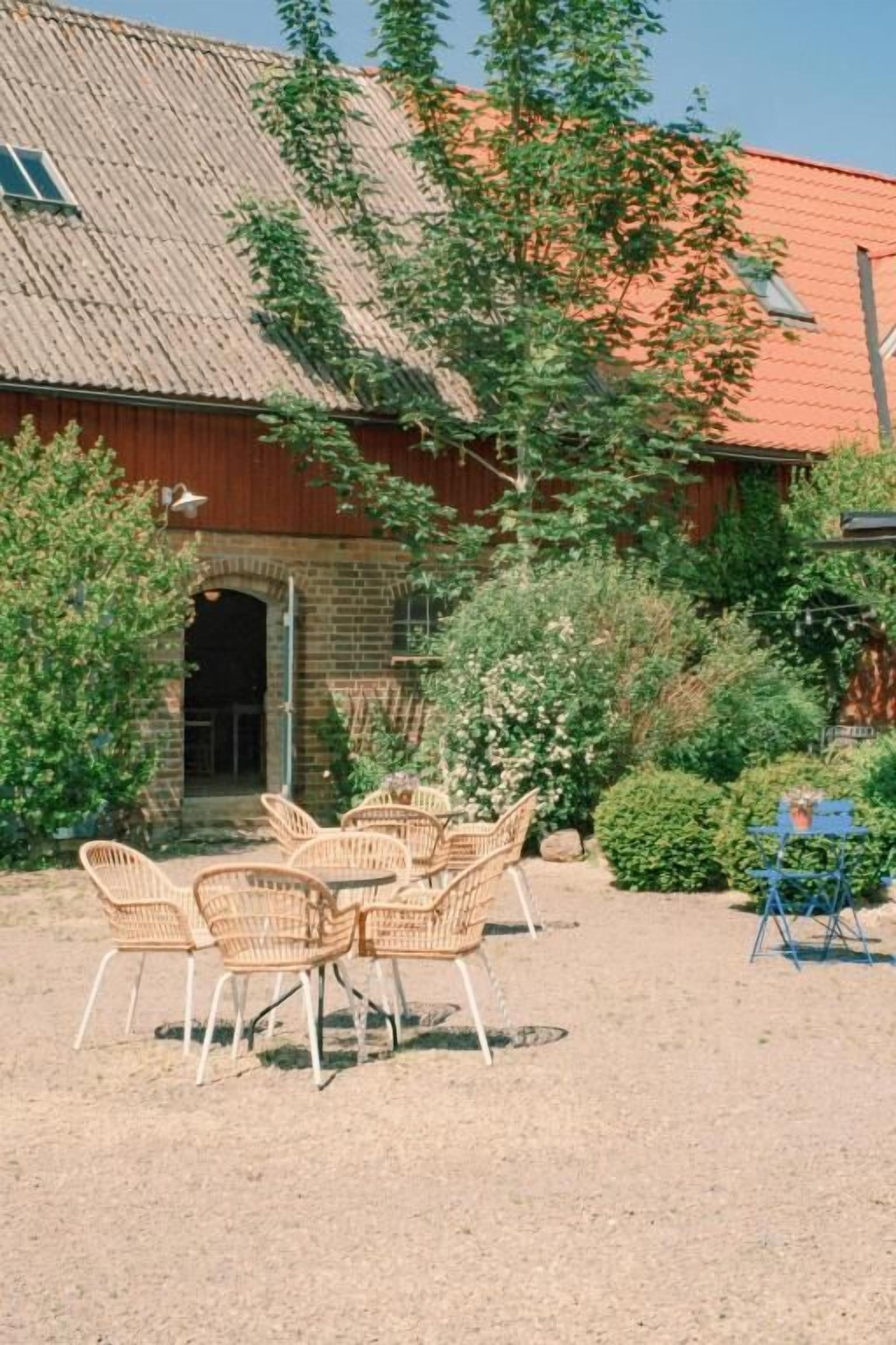 En byggnad med en fasad av tegel och ett tak täckt med takpannor, omgiven av grönska och ett område med grus och möbler av rotting.