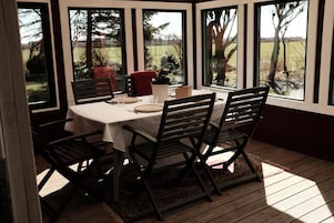 Uma área de refeições coberta com uma mesa posta para quatro pessoas, rodeada por cadeiras e grandes janelas que oferecem vista para o exterior.