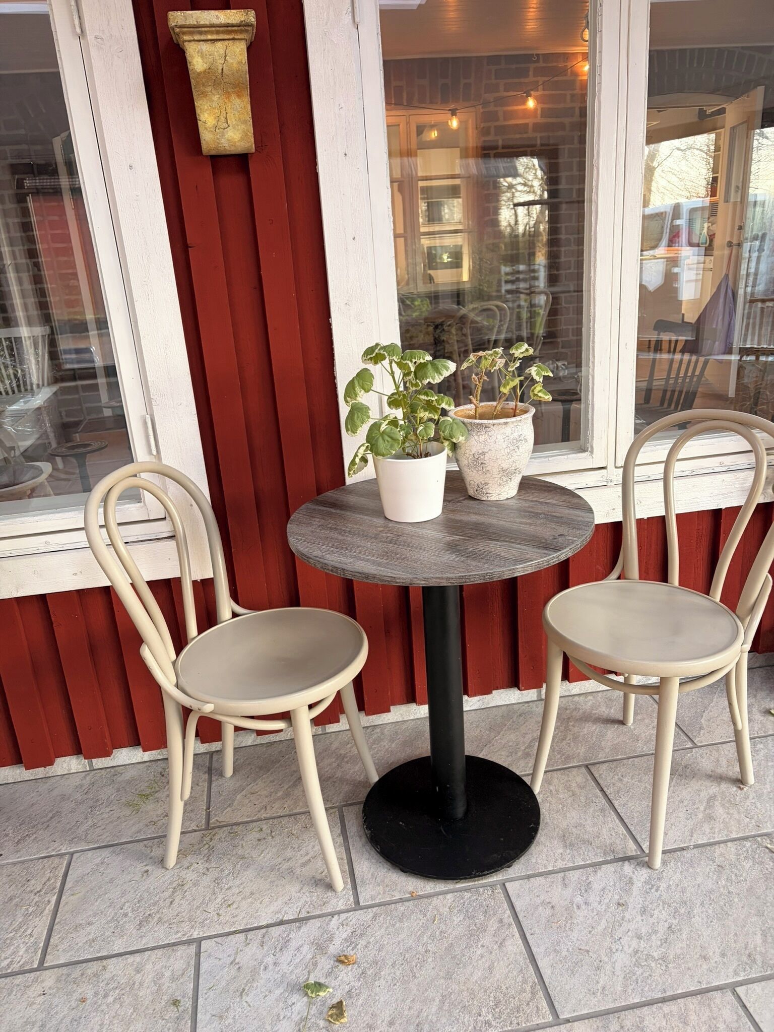 Una mesa redonda con dos sillas, dos plantas en macetas y una ventana que da al interior.