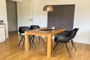 Un espace repas comprenant une table en bois, des chaises noires, un luminaire suspendu et un mur d’accent de couleur grise.