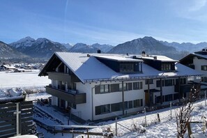 A modern building with a flat roof and large windows, surrounded by snow-covered ground and mountains in the background. - Holiday apartment with 2 bedrooms (Oberstdorf)