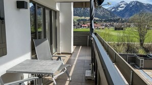 Een balkon met een tafel en stoelen, een uitzicht op een bergketen en een klein dorpje in de verte.