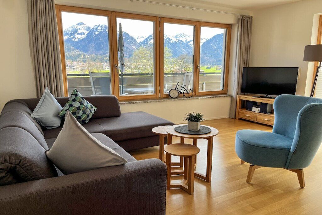 A living room with a sofa, a blue armchair, a small table with a plant, a television, and a window with a mountain view.