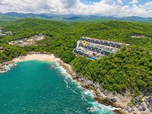 Ein Küstenresort mit einem großen Gebäudekomplex, umgeben von üppiger Vegetation, einem Sandstrand und türkisfarbenem Wasser.