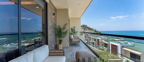 A balcony with a white sofa, potted plants, and a view of the ocean and buildings.