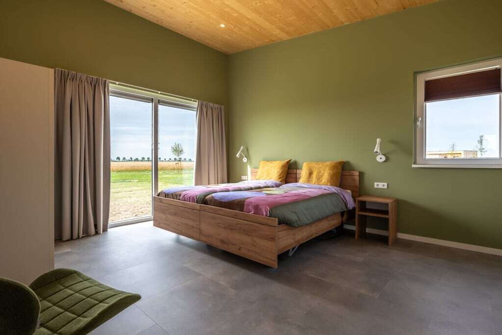 Un dormitorio con cama de madera, una mesita de noche pequeña, una silla y un ventanal con cortinas.