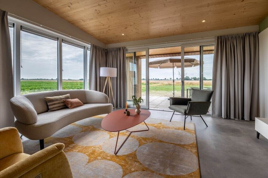 A modern living room with a sofa, armchair, and coffee table, large windows with curtains, and a view of the outdoors.