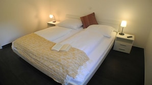 Un lit double recouvert d’une couverture rayée blanche et beige, deux tables de chevet avec des lampes et un oreiller rouge.