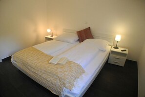 Een tweepersoonsbed met een wit-beige gestreept deken, twee nachtkastjes met lampen en een rood kussen.