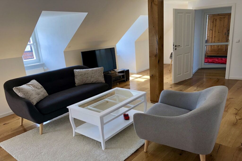 A modern living room with a black sofa, a white coffee table, and a grey armchair. There is a television on a stand and a wooden door leading to another room.