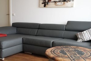 Ein graues Ecksofa mit einem roten Zierkissen und einem gestreiften Kissen, ein runder Couchtisch aus Holz mit Vintage-Design und ein Gemälde an der Wand.
