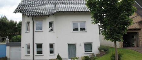 A two-story residential building with a tiled roof, multiple windows, and a small front yard with a tree.