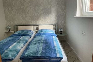 A bedroom with two single beds, a nightstand with a lamp, a small table with a lamp, and a window with a curtain. - Holiday apartment Rhine-Ahr (Sinzig)