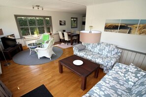 Ein Wohnzimmer mit einer Couch, einem Couchtisch, einem Esstisch mit Stühlen, einem Fernseher und einem großen Fenster mit Blick auf Grünflächen.