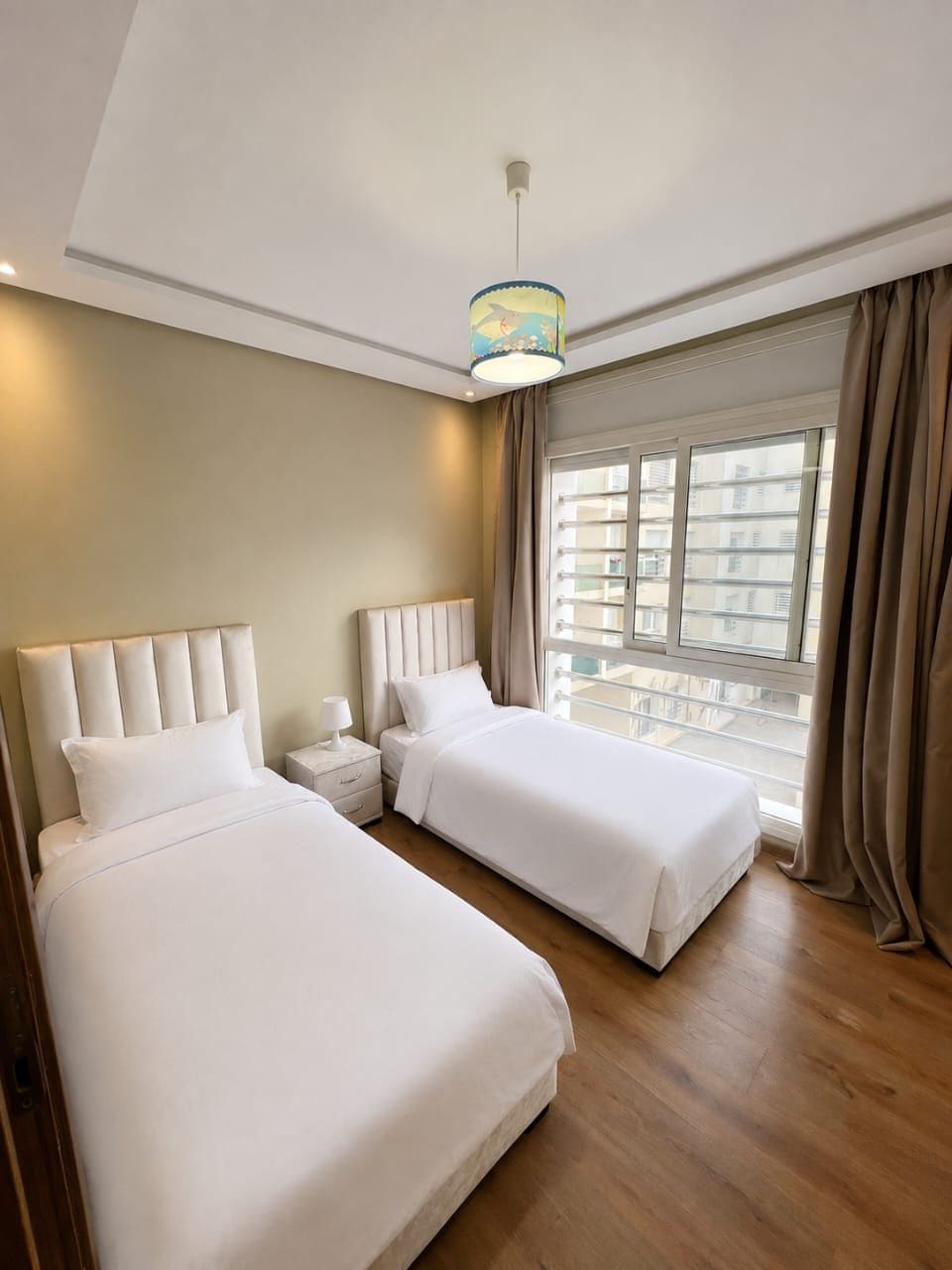 A hotel room with two beds, a window with curtains, a bedside table with a lamp, and a hanging light fixture.