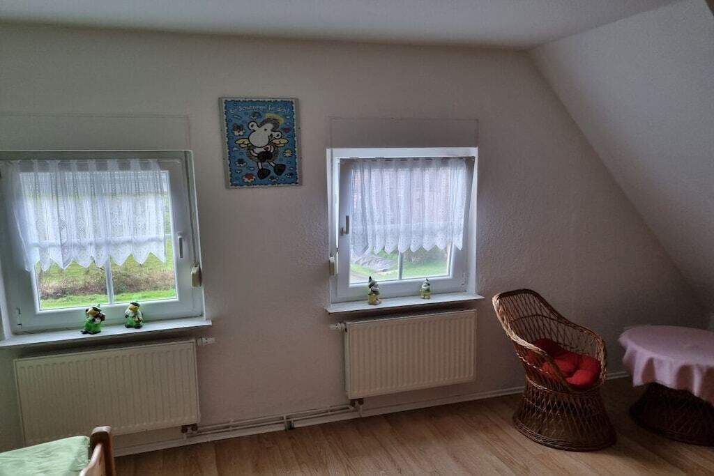 Een kamer met twee ramen, een radiator, een rieten stoel en een klein tafeltje.