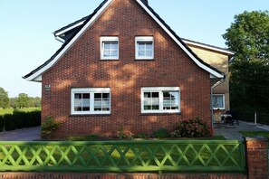 Een tweelaags bakstenen huis met een puntig dak, een groene omheining en een kleine tuin.