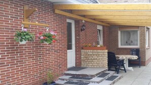 Un edificio de ladrillo con una pérgola de madera, un pequeño espacio al aire libre con dos sillas y una mesa, y plantas en macetas en la pared.