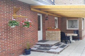 Een gebouw van baksteen met een houten pergola, een klein buitenterrein met twee stoelen en een tafel, en potplanten aan de muur.