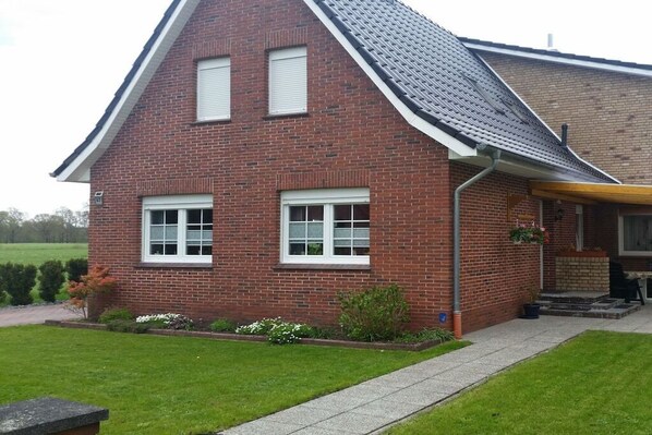 Een huis van rode bakstenen met een puntgevel, twee ramen met luiken, een klein portaaltje met een stoel, en een verzorgd gazon.
