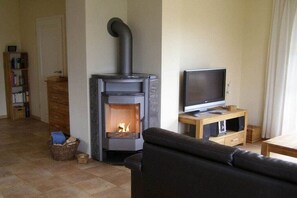 Ein Wohnzimmer mit Kaminofen, Fernseher auf Holzfuß, einer Couch und einem Bücherregal.