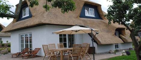 Une maison à toit de chaume dotée d’un patio, de mobilier d’extérieur et d’un grand parasol.